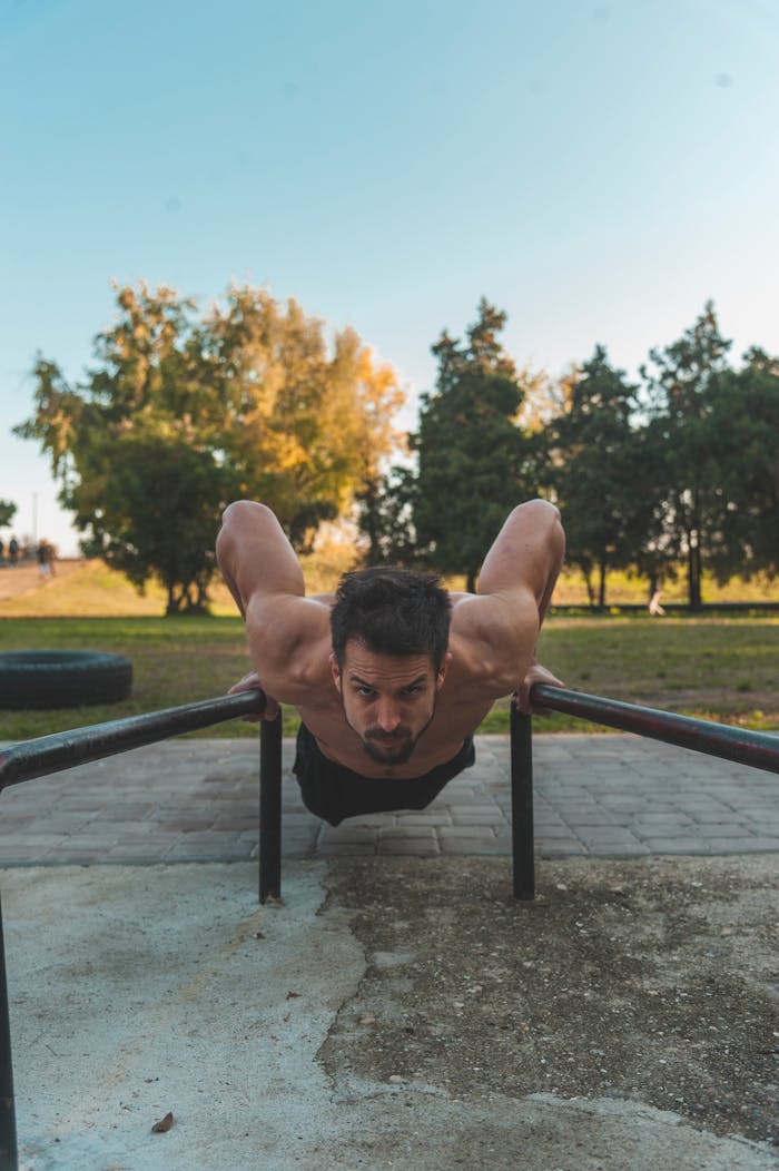 Home Shirtless man showcasing fitness by doing push-ups on metal bars in a park setting.